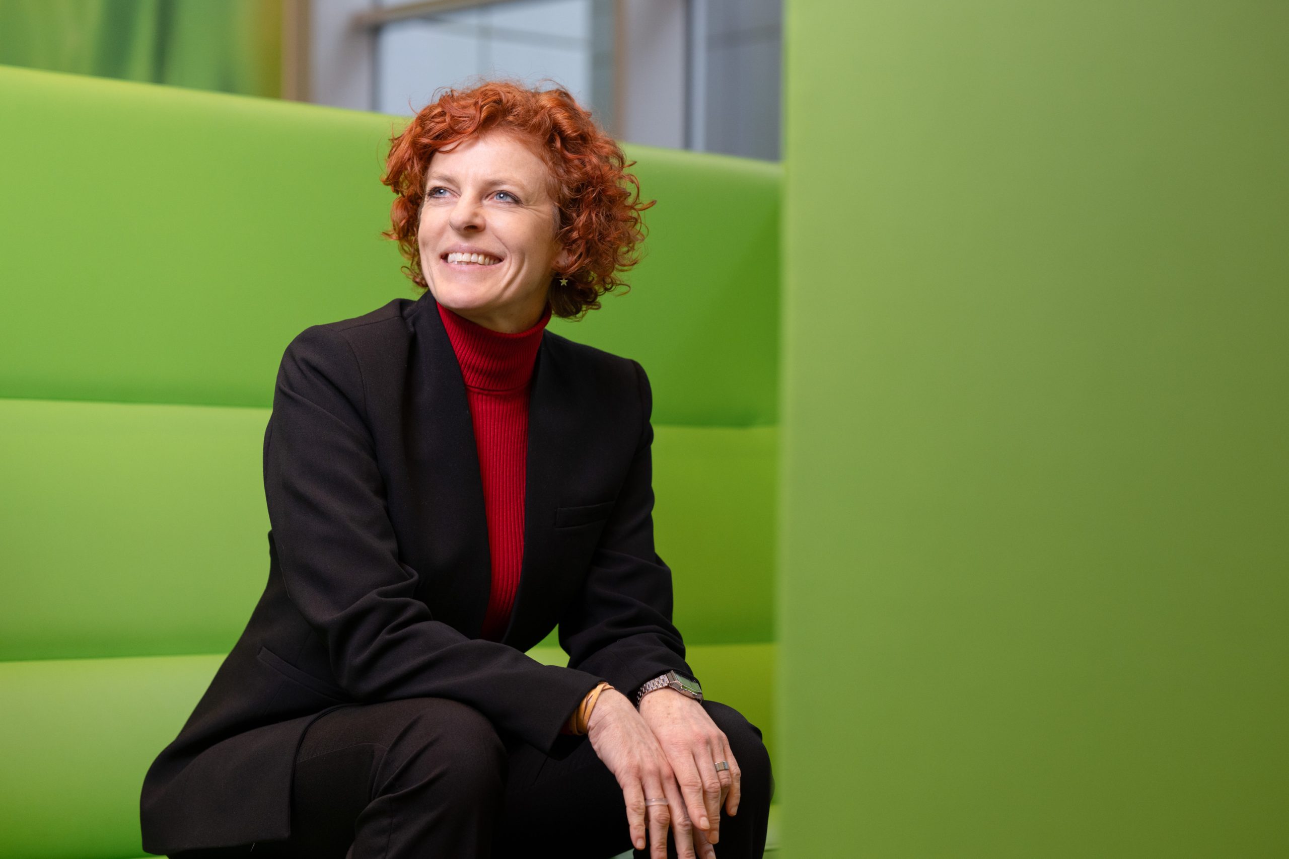 Marlies Klooster, vrouw met rood krullend haar, zwart mantelpak met rode coltrui eronder.